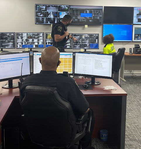 The image shows a security control room with several large monitors displaying various camera feeds